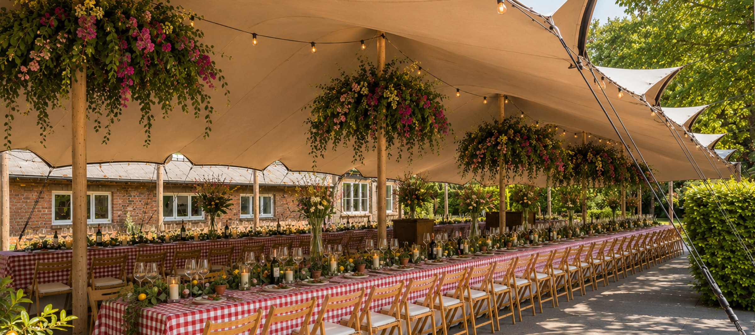 Lange gedekte tafels met rood-wit geblokte kleden onder een stretchtent, versierd met bloemen en lichtjes